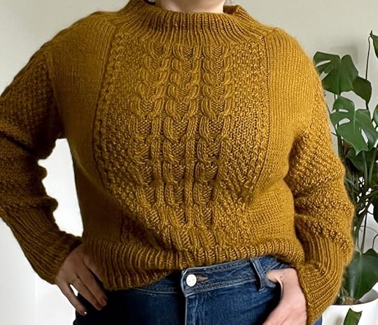 Close up of Shannon wearing a brown hand knit cabled sweater and blue jeans in front of white wall.