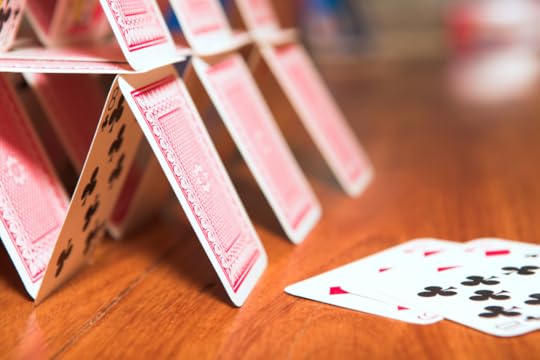 Image: a house made of precariously-stacked playing cards on a wooden table.
