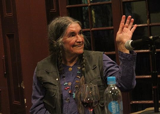 Bernadette Mayer sitting and waving before a microphone with a glass of wine and a water bottle.