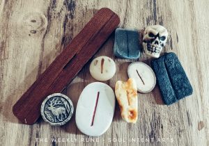 The elder futhark rune Isa carved into different media and pictured with a small plastic human skull and a reindeer carved into a metal button on a woodgrain background.