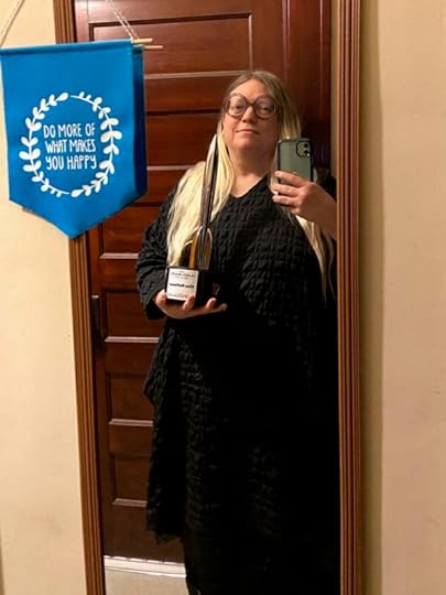 Elise, who is white with long blonde hair, wears a dark black dress. Elise is standing in front of a wooden door, holding up a phone camera to take the picture in one hand and the Hugo Award (shaped like a rocket ship) in the other. A blue hanging flag reads