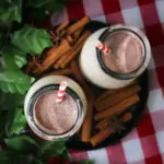 two glass bottles of milk nog surrounded by cinnamon sticks on a red checked cloth.