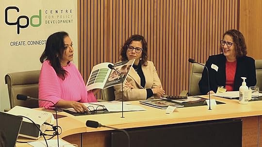 Three women at a conference table speaking into microphones and holding up a document