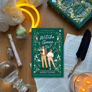 Book cover in front of a glowing crescent moon, scattered loose pages, a quill, a candle, vials of magical looking stones, and white roses