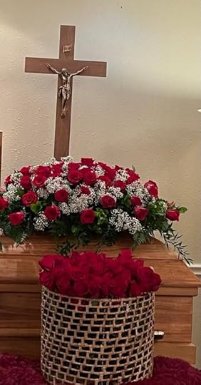 a large basket filled with dozens of red roses sits before a casket with a red rose spray, in front of a large crucifix.