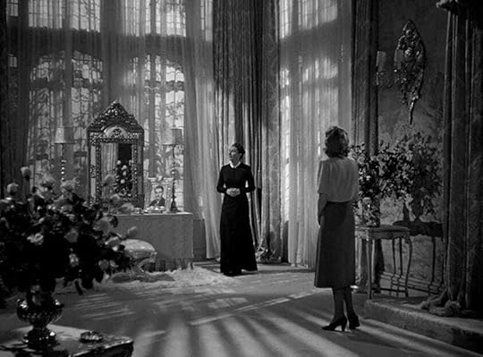 Judith Anderson as Mrs. Danvers in Rebecca (1940) stands in front of a draped window. Joan Fontaine as the second Mrs. de Winters looks at her.