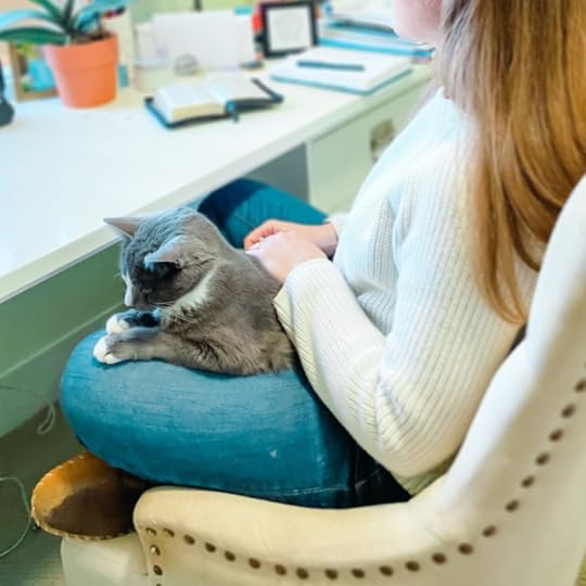 Amy Julia sits cross-legged at her desk and holds a gray cat in her lap