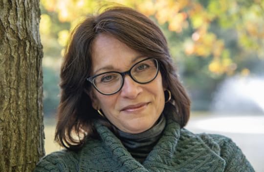 Color portrait photo of Meg Medina, smiling at camera. She has shoulder-length brown hair, frame glasses and is wearing a black turtleneck and a dark green sweater