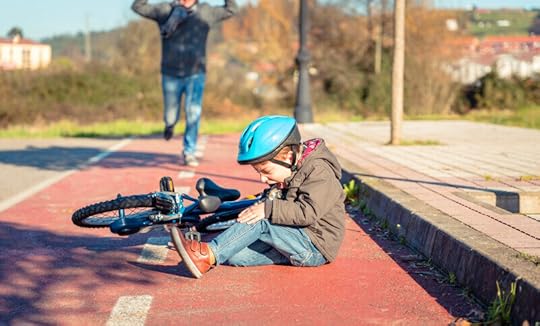 A little boy wearing a helmet sits on the ground at a park next to his toppled bicycle. He holds his knee and cries. A parent runs up to help. Dyspraxia can cause difficulty with motor skills, coordination, and balance.