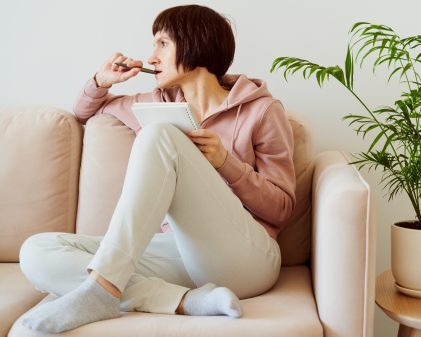Woman seated on sofa, staring off into the distance with a pen in her mouth, reflecting on something before she writes in the journal resting on her knee.