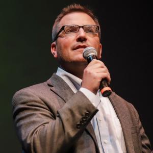 Author of 'CAPESTERS: A Hero Bureau Thriller' wearing a gray suit and glasses, holding a microphone speaking at an event.