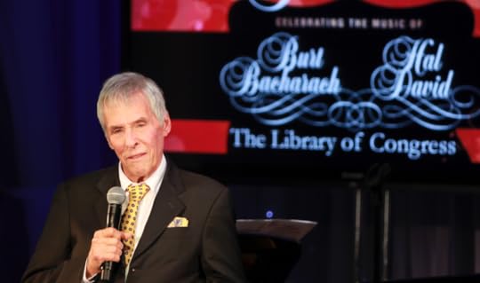 Burt Bacharach, in a suit and tie, holds a microphone while speaking on stage. A red screen in the background announces that he and lyricist Hal David are the 2012 Gershwin Prize honorees.