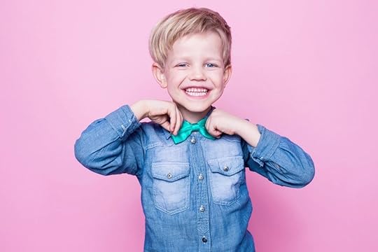 A beautiful young child with blonde hair smiles at the camera a big cheesy grin as they straighten up their bowtie.