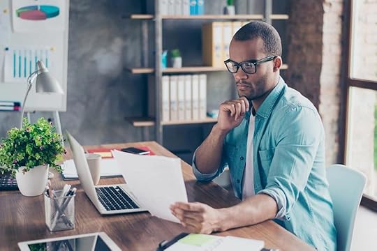 A man sitting at a desk holding a piece of paper with the other hand under their chin looking at it in concentration.