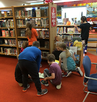 Group of fourth grade children gathered in library with hideous orange carpet, looking at books and searching the shelves.
