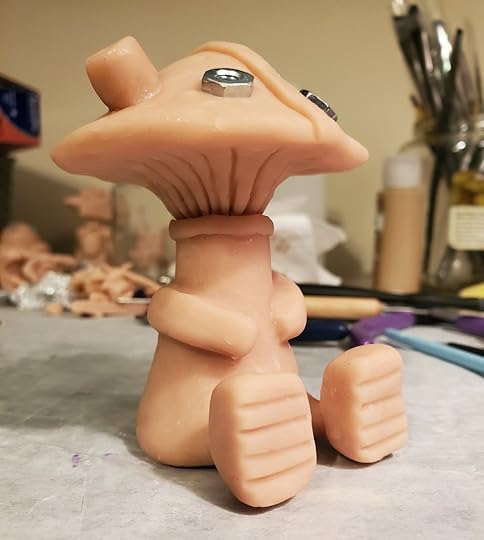 A finished sculpture of a mushroom robot. It isn't painted yet. It has tiny legs and arms and is sitting down on a messy craft table.