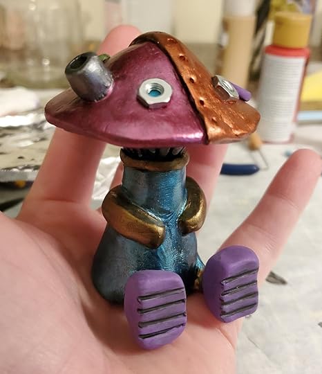 A small sculpture of a mushroom robot sitting in an open hand. The robot is painted with metallic paints of coper, bronze, teal, berry, and lavender. It has small metal nuts for eyes.