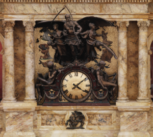 A wide view of the rotunda clock in the Library's main reading room, a sculpture of sybomlic figures (Father Time, etc) surrounding a large clock face. The statue and clock are flanked by columns