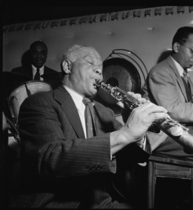 Black and white photo of Sydney Bechet, playing clarinet at a small jazz club. He's wearing a suit and tie and his eyes are closed in concentration