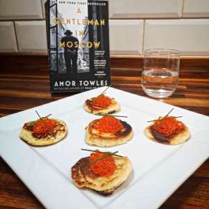 blinis topped wth caviar. In the background is the book A Gentleman In Moscow by Amor Towles and a glass of vodka.