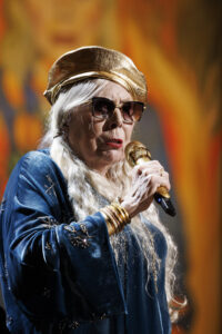 Medium close up of Joni Mitchell, holding a microphone, wearing dark glasses and singing on stage. The backdrop, a painting of hers, is bright orange.