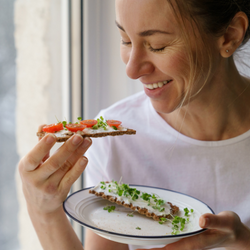 Woman holding a plate in one hand and a cracker with toppings in other hand. She is looking at it, smiling, and about to take a bite.