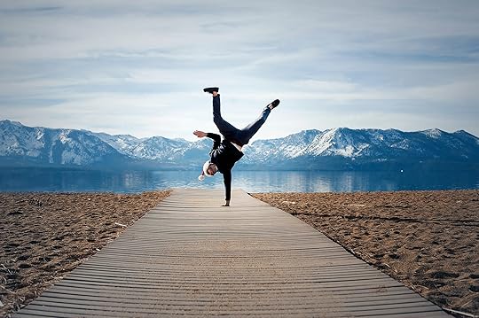 Person showing some creative confidence while doing a one-handed handstand on a dock leading to a blue lake.
