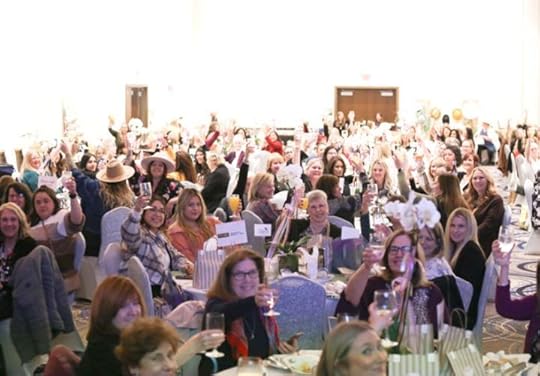 Many women sitting in a hotel ball room at tables holding up glasses in a toast.