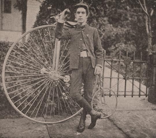 A young man stands next to a 19th century bicycle, with the front wheel almost as tall as he is and very small rear wheel.