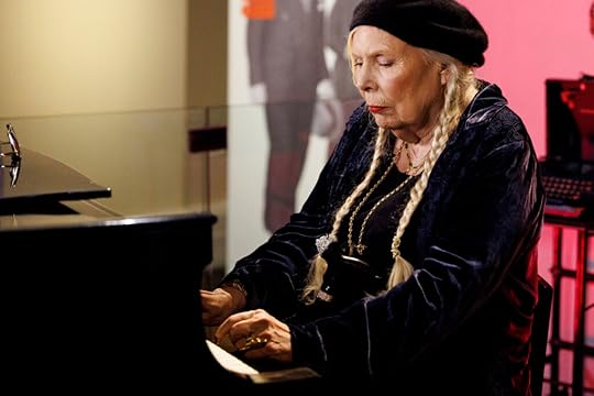 Joni Mitchell, wearing a beret, with long blond braided pigtails falling over her shoulders, sits at a black piano, hands on the keyboard.