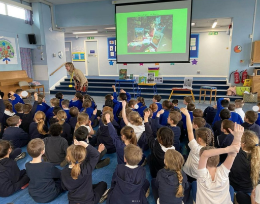 School children seated in a school hall with hands up during an author talk by Karen Inglis who is standing at the front of the class taking questions.