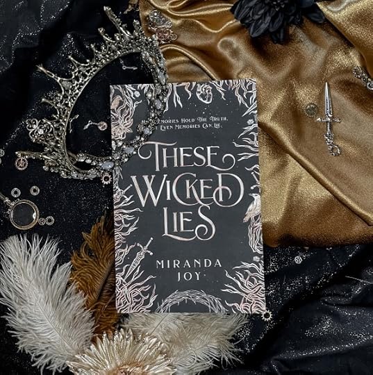 These Wicked Lies by Miranda Joy (a new adult fantasy romance book) is propped on gold and black fabric with feathers and a crown.