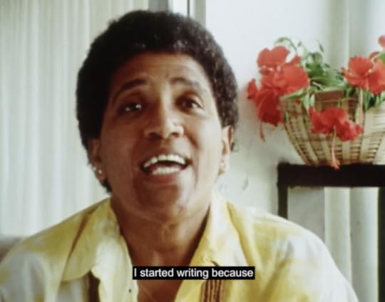 Audre Lorde sits wearing a yellow shirt in a white room with red flowers in the background. She begins, "I started writing because"