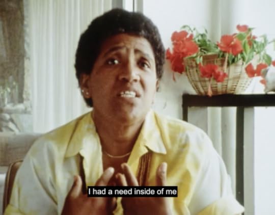 Audre Lorde sits wearing a yellow shirt in a white room with red flowers in the background. She holds her hands turned inward toward her chest and continues, "I had a need inside me"