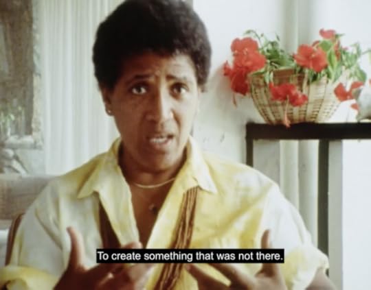 Audre Lorde sits wearing a yellow shirt in a white room with red flowers in the background. She turns her hands outward and concludes, "To create something that was not there."