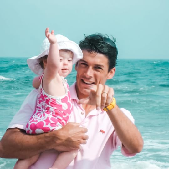 Peter holds a baby Penny and points at the camera while Penny raises her arm in the air. They are standing in front of the ocean.
