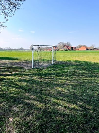 Ein Fußballtor steht auf einer weiten, leeren Rasenfläche, im Hintergrund ziegelrote Bauernhäuser