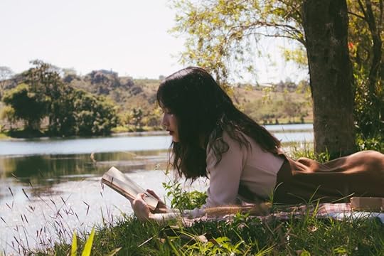 A young girl reading a book on the grass near a pond.