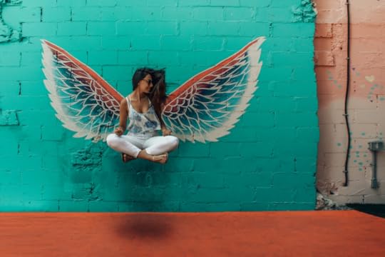 Image: a colorful pair of wings are painted on a cinderblock wall. Seeming to float midair in front of the wings as if they belong to her, a woman sits crosslegged.