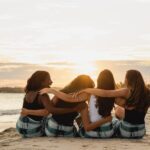 a group of four women with their backs to the camera arms around each other, sit on the beach watching the sunset in the distance.