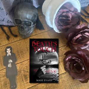 Picture of book cover on a wooden backdrop. A toppled white vase with dark purple roses is pouring down the right of the book, a black skull is on the top left, and a paper