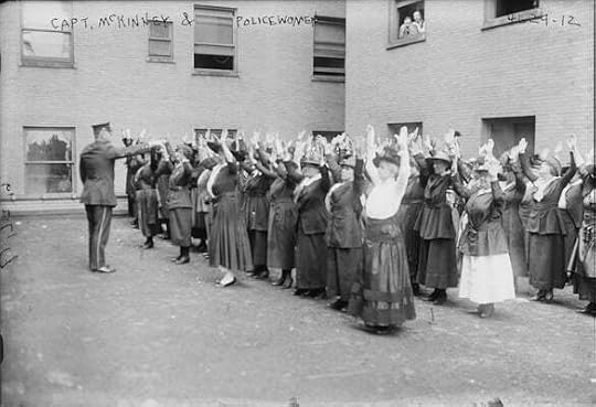 Black and white photograph of a crowd of women exercising in a courtyard under the supervision of a tall, white, male in police uniform. The image has a handwritten caption saying 