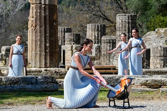 Standing in front of Ancient Greek ruins, a woman dressed in a white robe lights an Olympic torch in a parabolic mirror.