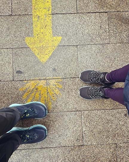 Two pairs of feet, one in black sneaks and the other in blue sneakers, stand next to a yellow arrow and seashell