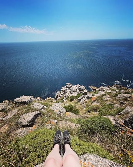 A pair of feet in black sneakers hangs out over the rock cliff, the dark blue ocean beyond