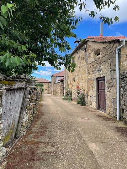An old paved road stretches forward between an old wooden fence leaning precariously and a tan-and-light-grey-stone house with a dark wooden door and plants growing out of the cracks