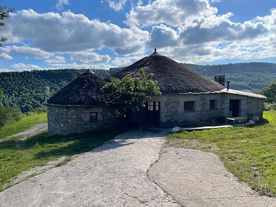 A circular, stone building with a thatched roof, rolling green pastures in the distance with a bright blue sky and fluffy clouds