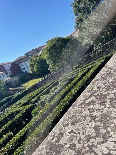 A dark green labyrinth hedge sits in a stone-walled courtyard with white and red buildings in the background
