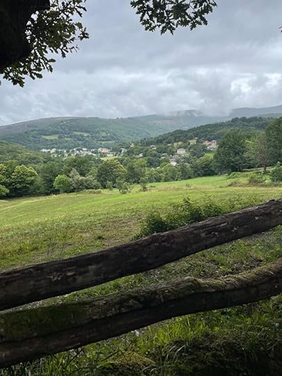Old wooden fence sits before a green pasture, and in the distance, pale buildings scattered between dark trees with a larger mountain in the background and a thick cover of clouds