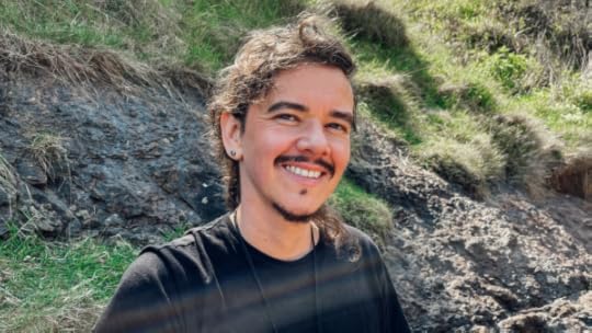 Photo shows author Die Booth smiling in front of a grassy cliff background.